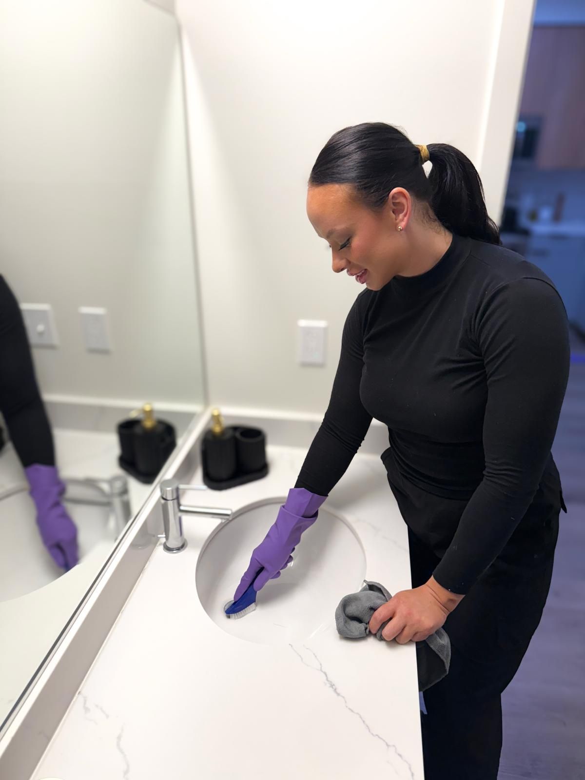 A professional cleaner wearing gloves scrubs a bathroom sink with a brush while holding a cloth, demonstrating detailed cleaning of surfaces and fixtures in a modern, well-maintained residential bathroom space.