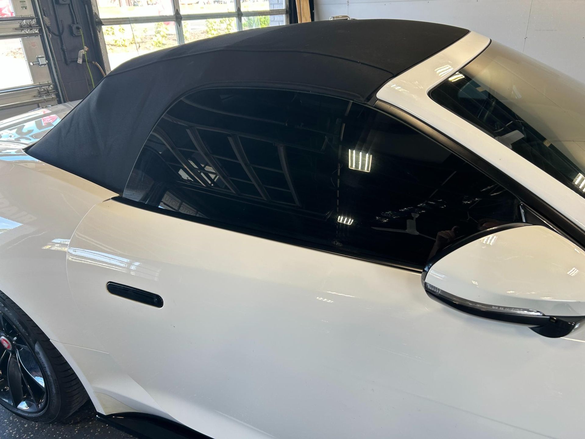 A close-up view of a sleek, white convertible sports car with a black fabric roof and tinted windows. The image highlights the car's clean finish and professional window tint installation in a garage setting.