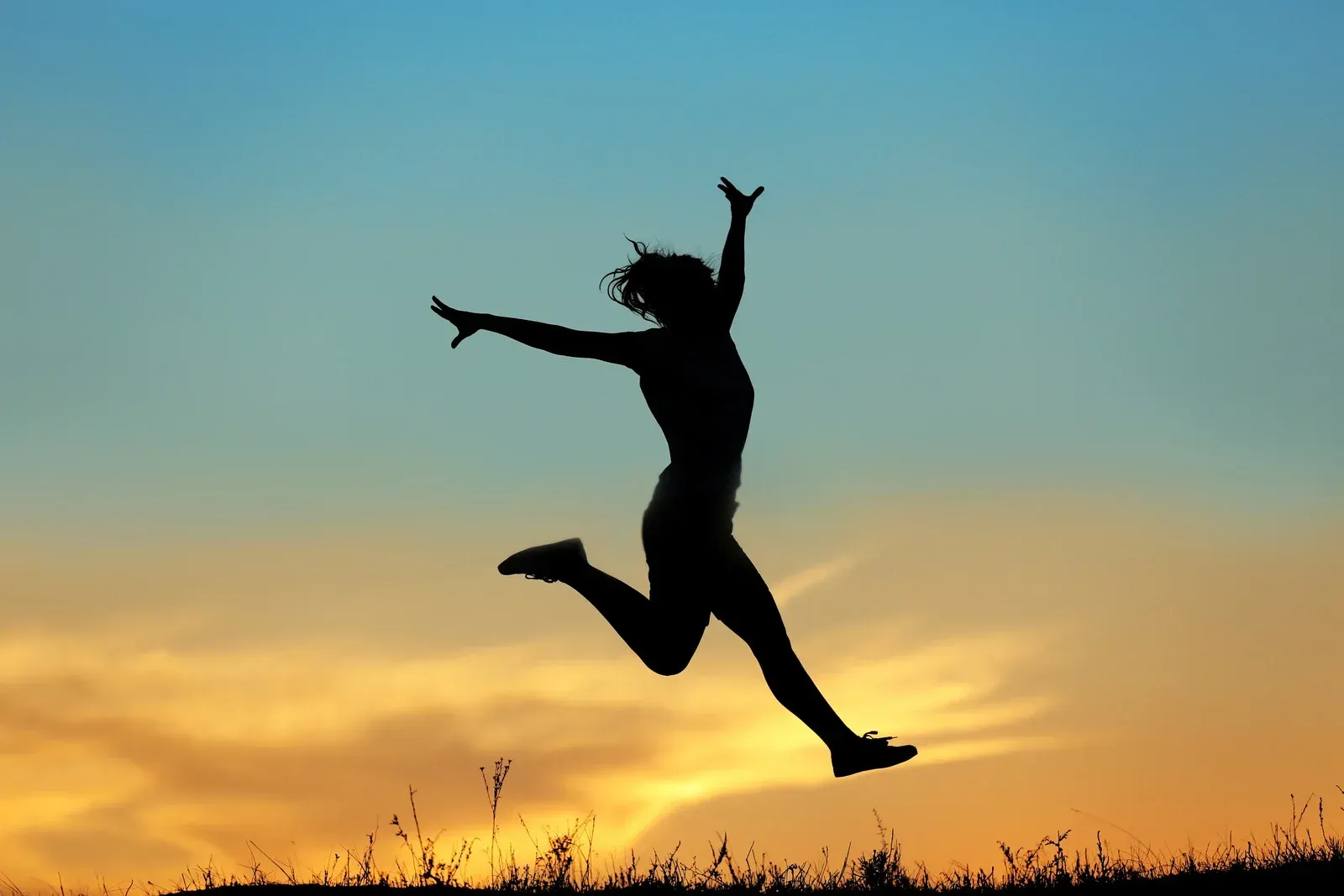 A dark silhouette of a person is captured mid-jump against a vibrant gradient sky. The sky transitions from a deep blue at the top to warm orange and yellow hues near the horizon, suggesting either sunrise or sunset.