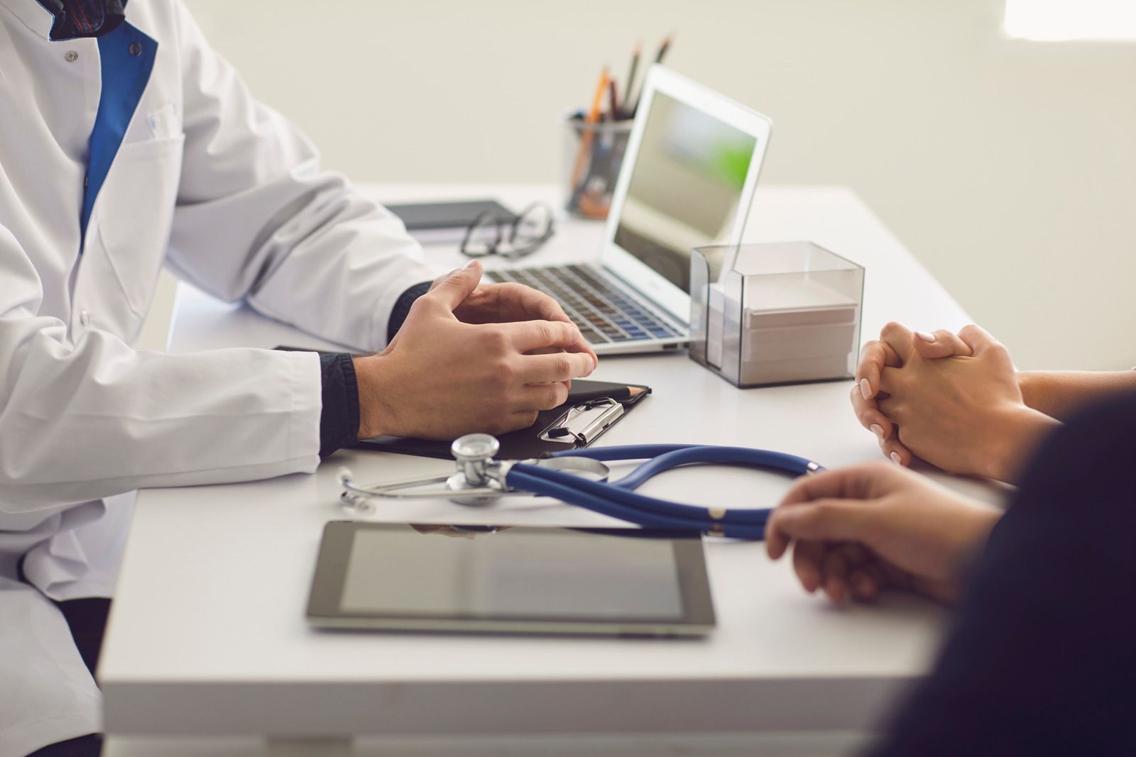 A doctor in a white coat sits at a desk discussing health information with a patient. A stethoscope, tablet, clipboard, and laptop are visible, indicating a professional medical consultation.