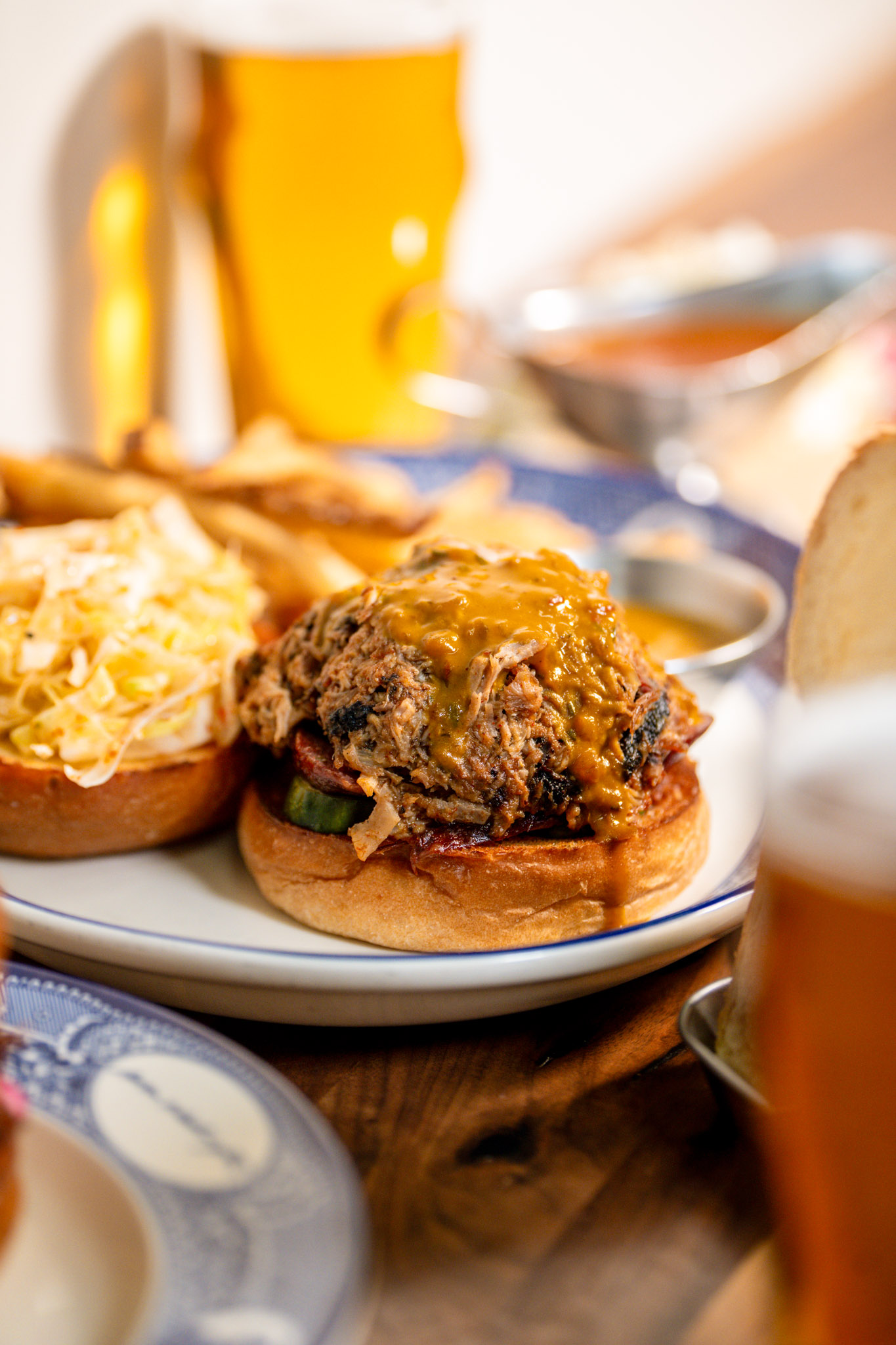 Carolina Pulled Pork & Sausage Sandwich on a toasted potato roll, draped in carolina gold mustard BBQ sauce with bread and butter pickles and spicy pickled cabbage, served alongside fries and coleslaw, with cold beers in the background.