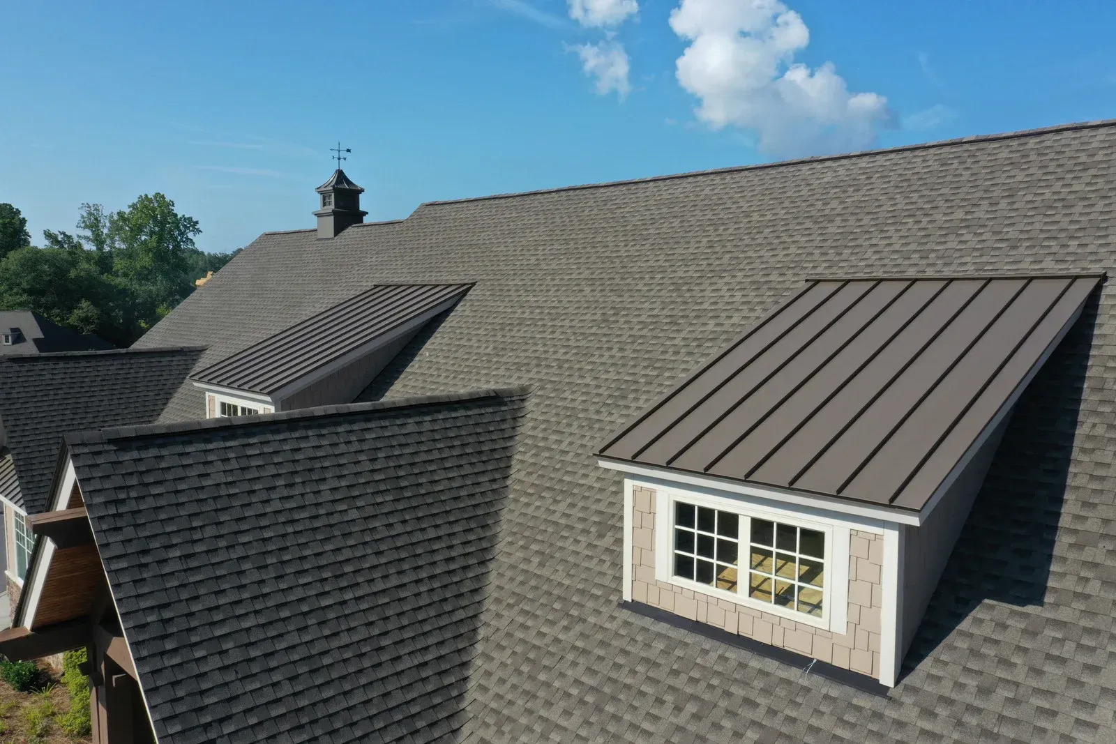 Large residential roof featuring asphalt shingles and standing seam metal dormers. The design combines traditional roofing materials with modern accents to enhance durability and visual interest.
