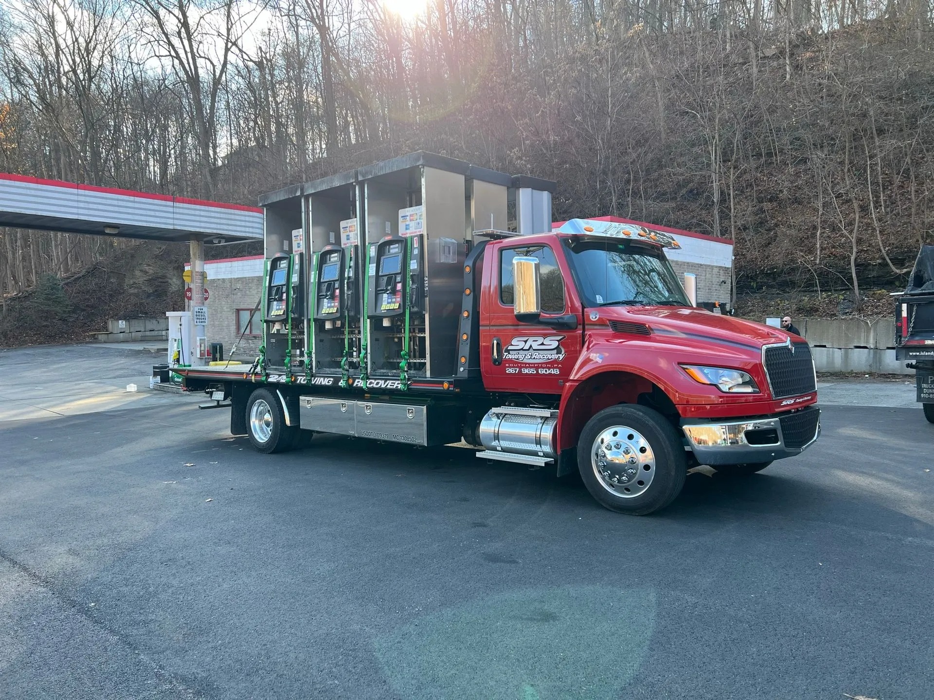A red SRS Towing & Recovery flatbed tow truck carrying multiple fuel dispensers, securely strapped, parked at an empty gas station near a wooded hillside.