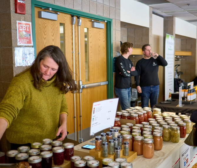 A female vendor in a green sweater organizing rows of glass mason jars containing home-canned jams, salsas, pickles, and spices on a tabletop. Signs in the background indicate EBT/SNAP payment acceptance at the indoor community center venue