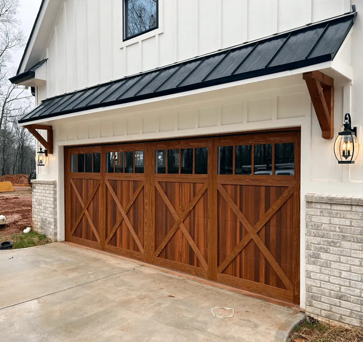 A custom carriage-house style garage door combines rich wood tones, decorative crossbuck detailing, upper glass panels, exterior lanterns, and white siding for a classic architectural appearance with rustic elegance and charm.