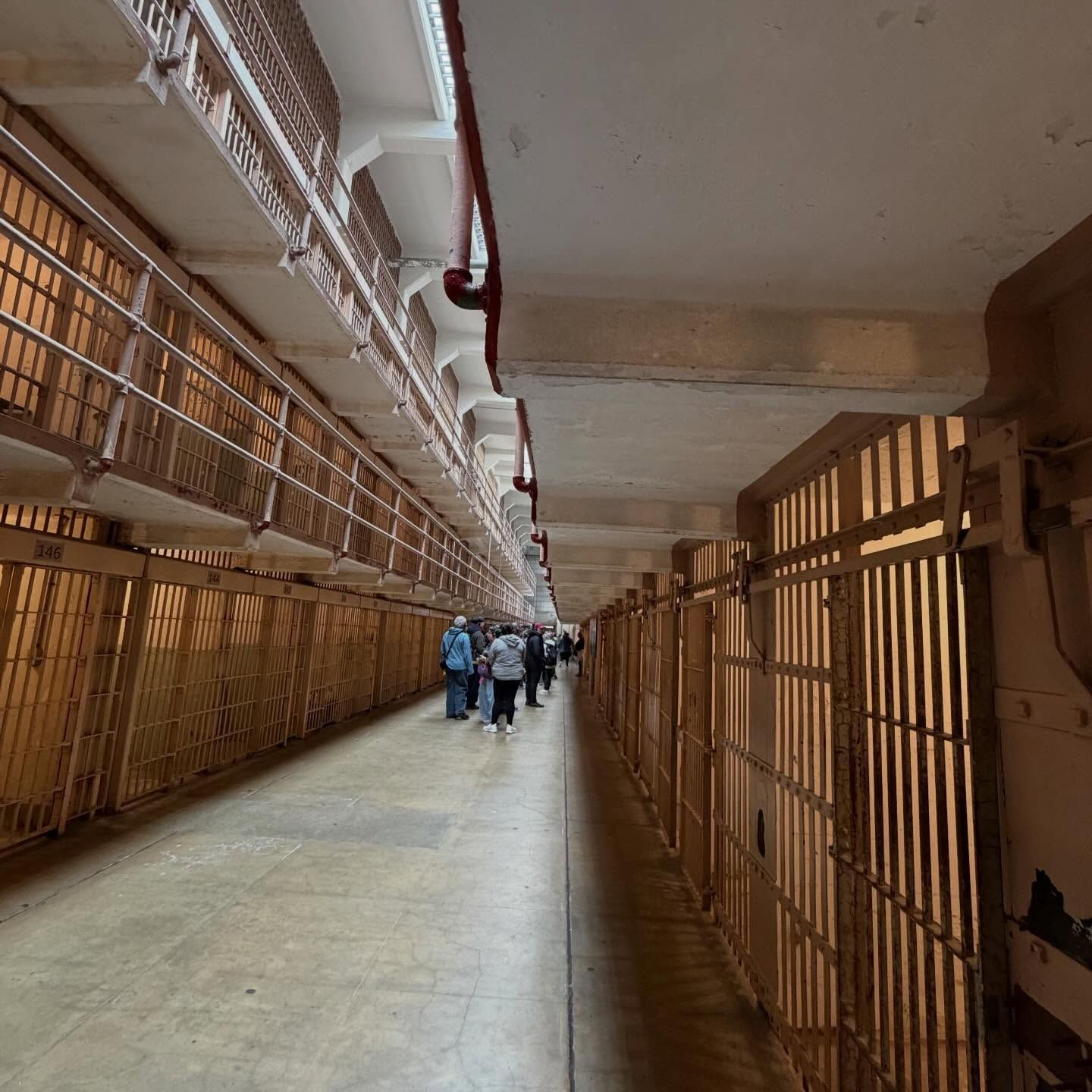 Long prison corridor lined with metal-barred cells on both sides, featuring multiple tiers and a group of visitors walking through, highlighting institutional architecture, confinement design, and historical correctional facility environment.