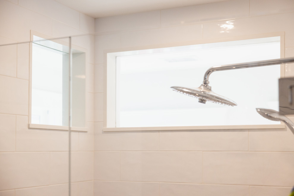 A close-up shot of a modern bathroom shower with white tiled walls, two chrome shower heads, and a narrow window. The lighting is bright and airy, creating a clean and minimalist aesthetic.