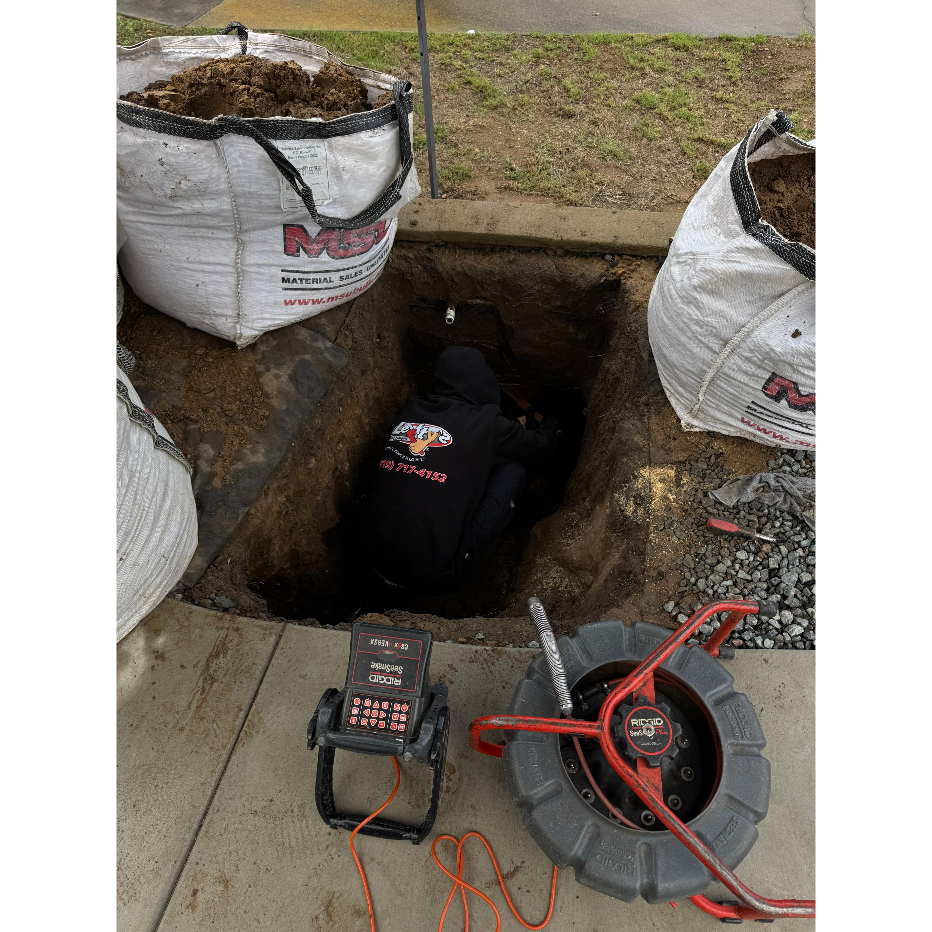 The technician is deep in the trench, working diligently to fix a plumbing issue. The image also shows the equipment used, including a camera system for inspections, and bags of soil nearby, illustrating the work required for a thorough repair.