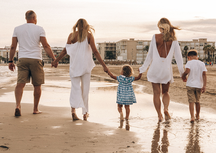 A family of five walking hand-in-hand along a sandy beach at golden hour, viewed from behind — two parents in white clothing guide three young children near the shoreline, with a coastal skyline in the distance, evoking generational wealth planning and long-term family financial security.