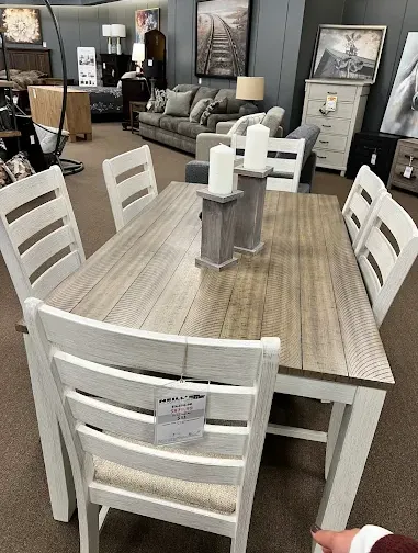 Dining room furniture display featuring a rectangular wood dining table with matching chairs, decorative candle holders, and nearby living room and accent furniture.