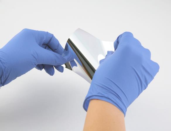 Technician in blue nitrile gloves handling DSI's Sunshade® Tape flexible optical solar reflector (FOSR) material. This innovative product offers equivalent optical properties to traditional glass optical solar reflectors (OSRs) with dramatically reduced manufacturing costs, easier installation via 3M966 pressure-sensitive adhesive backing, and superior durability during handling. Ideal for satellite radomes, antenna covers, and apertures requiring thermal control with ESD protection.