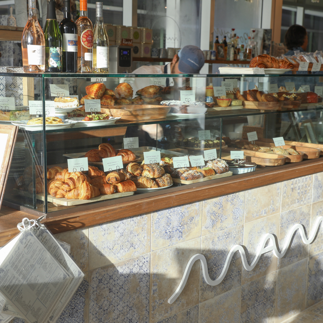 Morning light falls across the pastry counter, where rows of freshly baked pastries, cakes, and savory dishes are displayed beneath glass alongside bottles of wine and café details.
