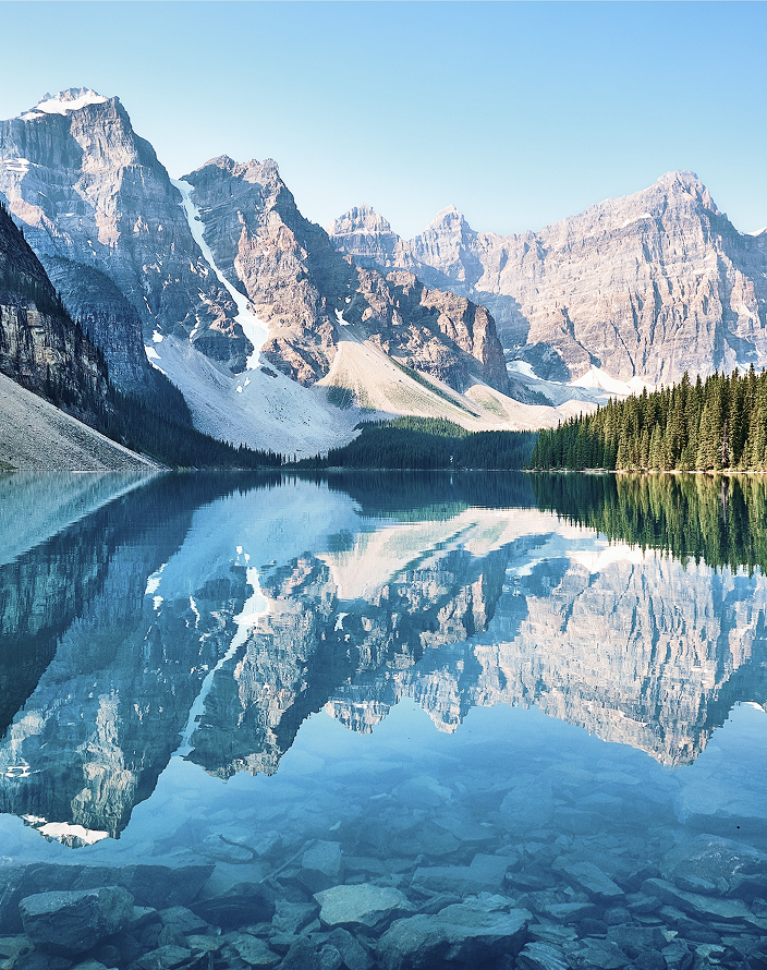 A calm, mirror-like alpine lake surrounded by rugged, snowy mountains and evergreen trees on a sunny winter day.