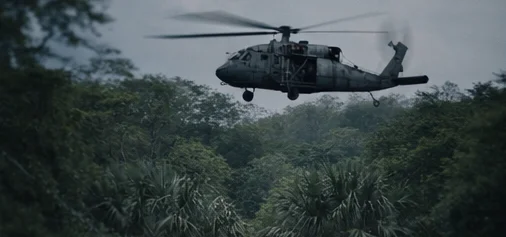 A military helicopter is captured in mid-flight above a dense, tropical forest under a cloudy sky. The helicopter's side door is open, and its rotor blades are blurred, indicating motion. The surrounding foliage is lush and green, suggesting a remote, natural environment.