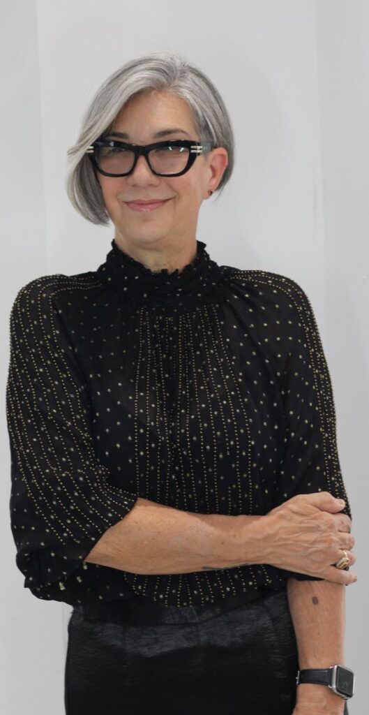 A medium shot of a woman with short, gray hair and glasses, wearing a black blouse with a gold dotted pattern and a black skirt. She is standing against a white wall with her arms crossed.
