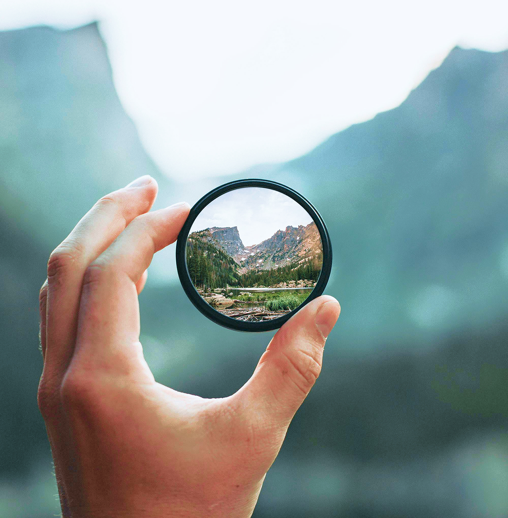 A hand holding a mirror that reflects a scene with mountains, trees, and a lake.
