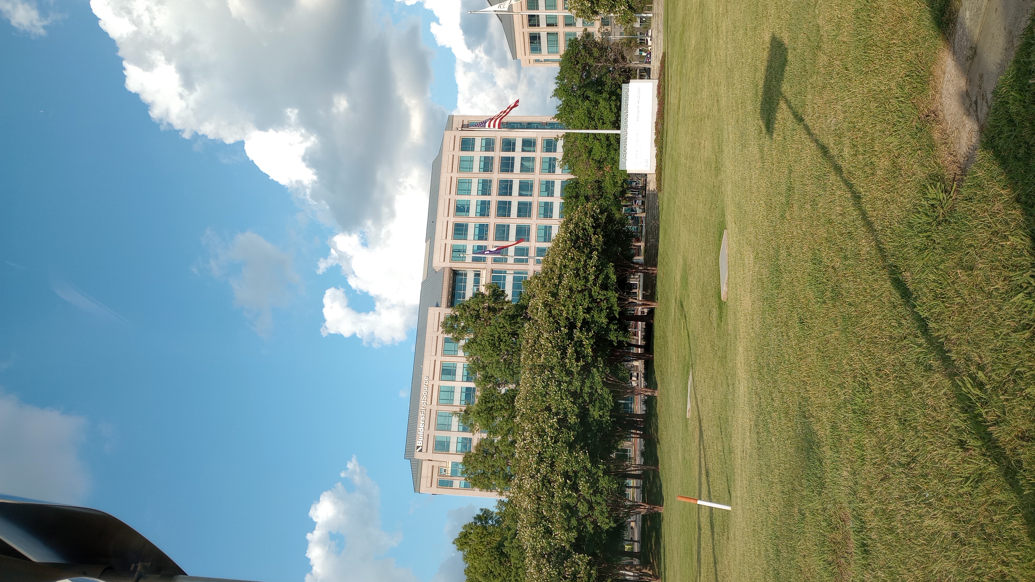 The image shows the exterior of a large, multi-story office building labeled "Builders FirstSource" at the top. The building has a modern design with large glass windows and is surrounded by well-maintained landscaping, including a row of trees in the foreground. Two flags, one American and one likely the Texas state flag, are flying on flagpoles near the building. The sky is mostly clear with some scattered clouds, and the sun is shining brightly, casting shadows on the grassy area in front of the building