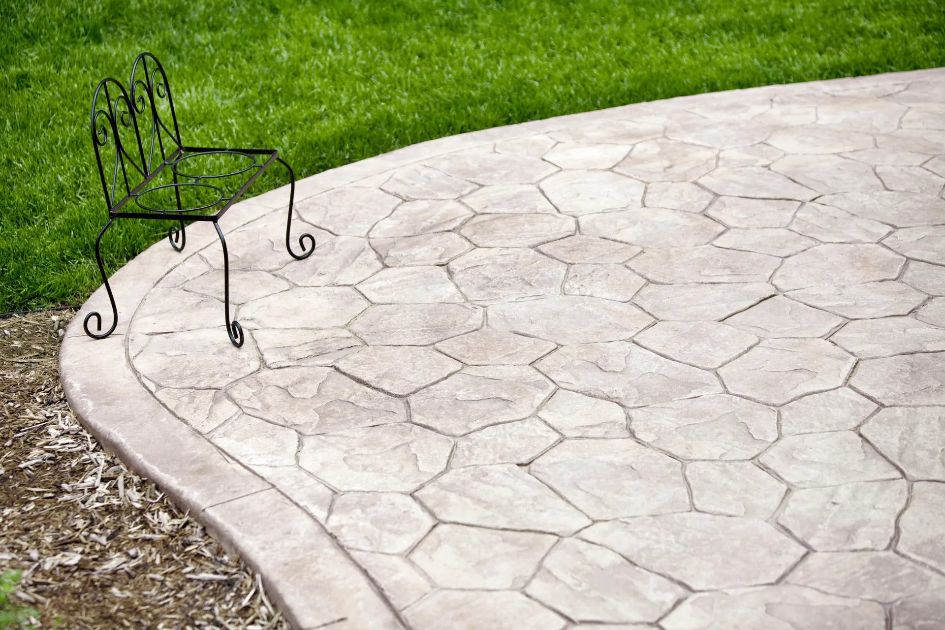 A decorative black metal stand with scrolled legs sits on a stamped concrete patio next to a neatly trimmed green lawn.