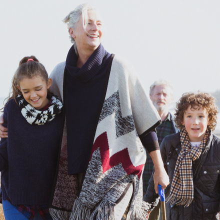 A joyful family strolls along a grassy beach path, with the ocean in the background. A young girl holds a fishing net, smiling alongside an older woman.