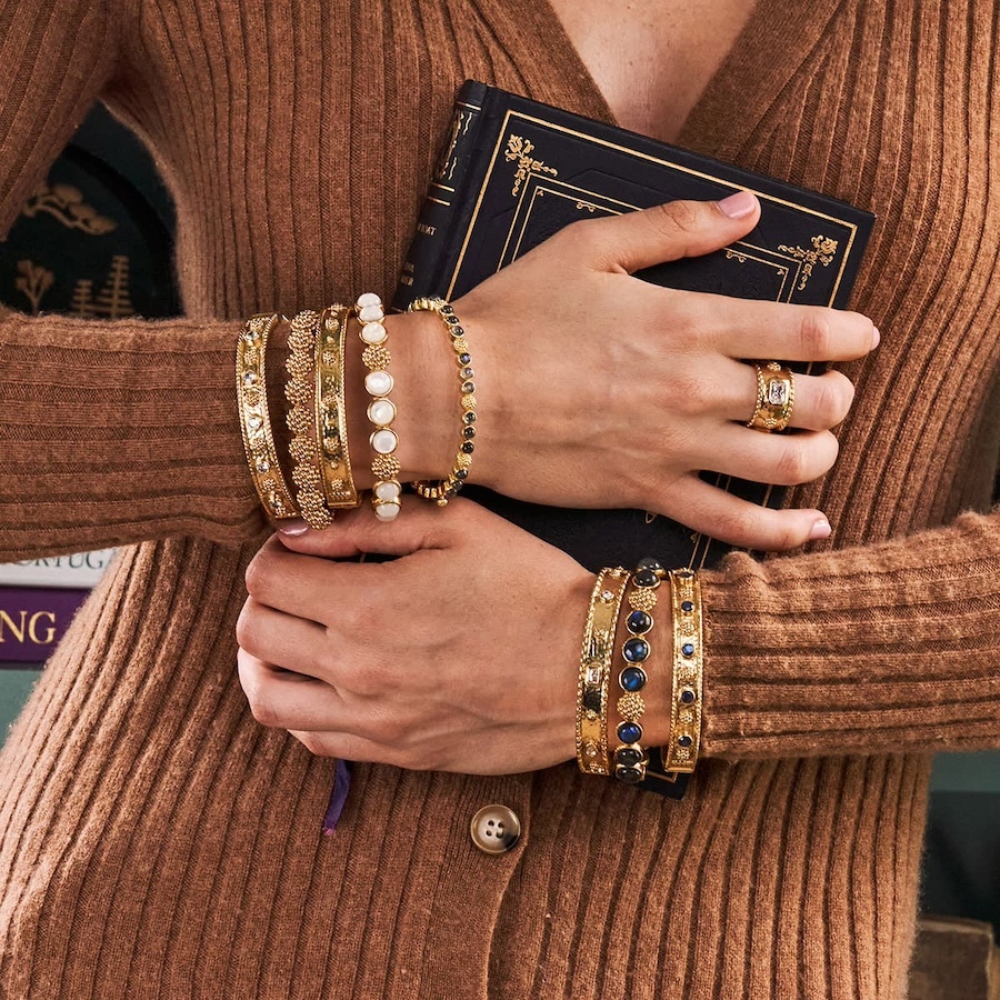 A compelling visual by Tabula Rasa Social Stationers showcasing the artistry of Capucine De Wulf jewelry. The image features hands adorned with a curated stack of gold-toned bracelets, some intricately detailed with pearls and others with deep blue gemstones, alongside a complementary ring. This collection embodies a blend of bohemian elegance and modern sophistication, crafted with intention from ethically sourced materials and inspired by nature's organic forms, reflecting the timeless and uniquely personal appeal of Capucine De Wulf designs.