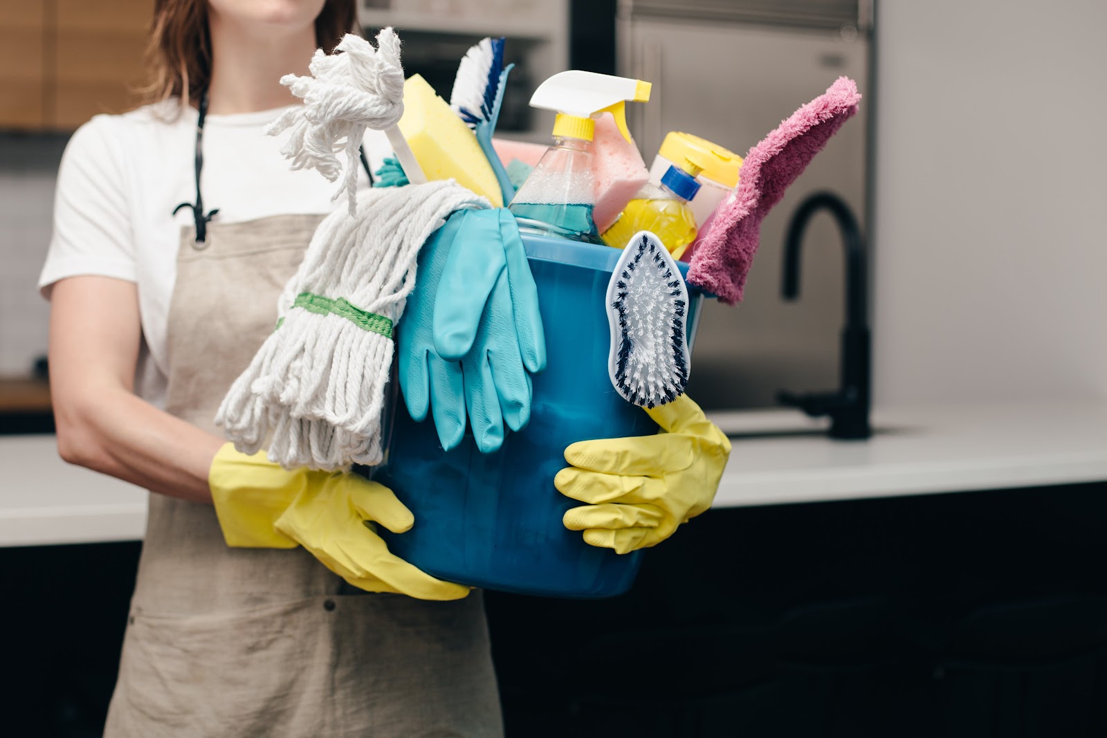 A person wearing yellow cleaning gloves and a beige apron is holding a blue bucket filled with various cleaning supplies, including mops, sponges, brushes, and spray bottles. The background appears to be a modern kitchen or bathroom with a sink and cabinets.