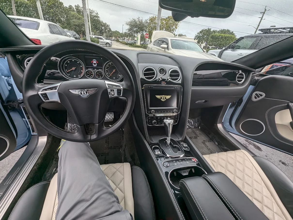 A person's legs are visible in the driver's seat of a luxury car. The car's interior is visible, including the steering wheel with the Bentley logo, the dashboard with a digital display and infotainment screen, and the center console with a gear shifter and cup holders. The seats are made of beige and black leather with diamond stitching. The exterior of other cars and a tree-lined street are visible through the car's windows.