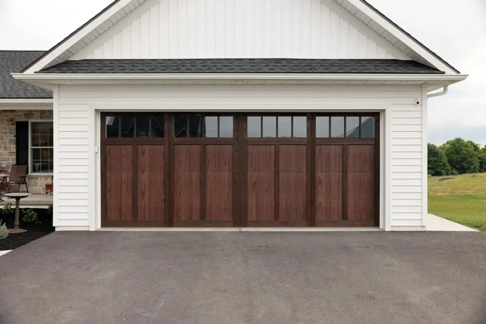 A residential garage features a warm wood-look double garage door with upper windows, white siding, dark shingles, and a clean paved driveway creating an inviting exterior with classic curb appeal.