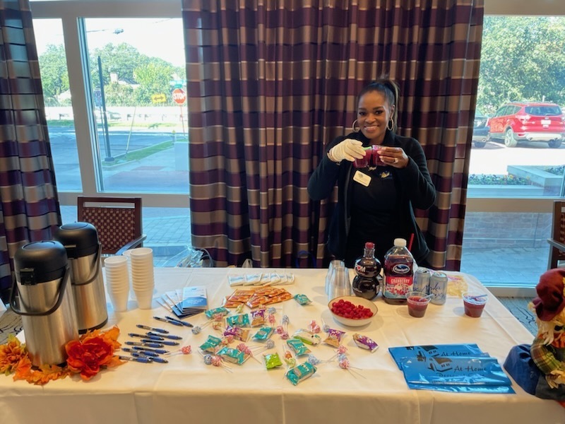 A woman in a black jacket and white gloves is standing behind a table filled with various items. She is holding up two small cups of a red beverage, seemingly offering them. On the table, there are two large silver thermoses, stacks of white paper cups, assorted candies and lollipops, pens, a bottle of what appears to be cranberry juice, another bottle with a red label, cans, a bowl of raspberries, and some brochures or flyers. The scene is set indoors with large windows in the background, revealing trees and a street outside. Decorative fall foliage and a scarecrow figure are also visible on the right side of the table.