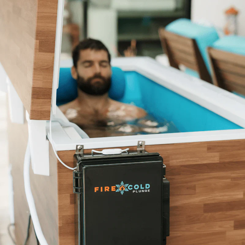 A man with a beard is submerged in a cold plunge tub, his eyes closed and head resting on a blue pillow. The tub has a wooden exterior and a blue interior filled with water. A black control box with the "FIRE & COLD PLUNGE" logo is attached to the front of the tub.