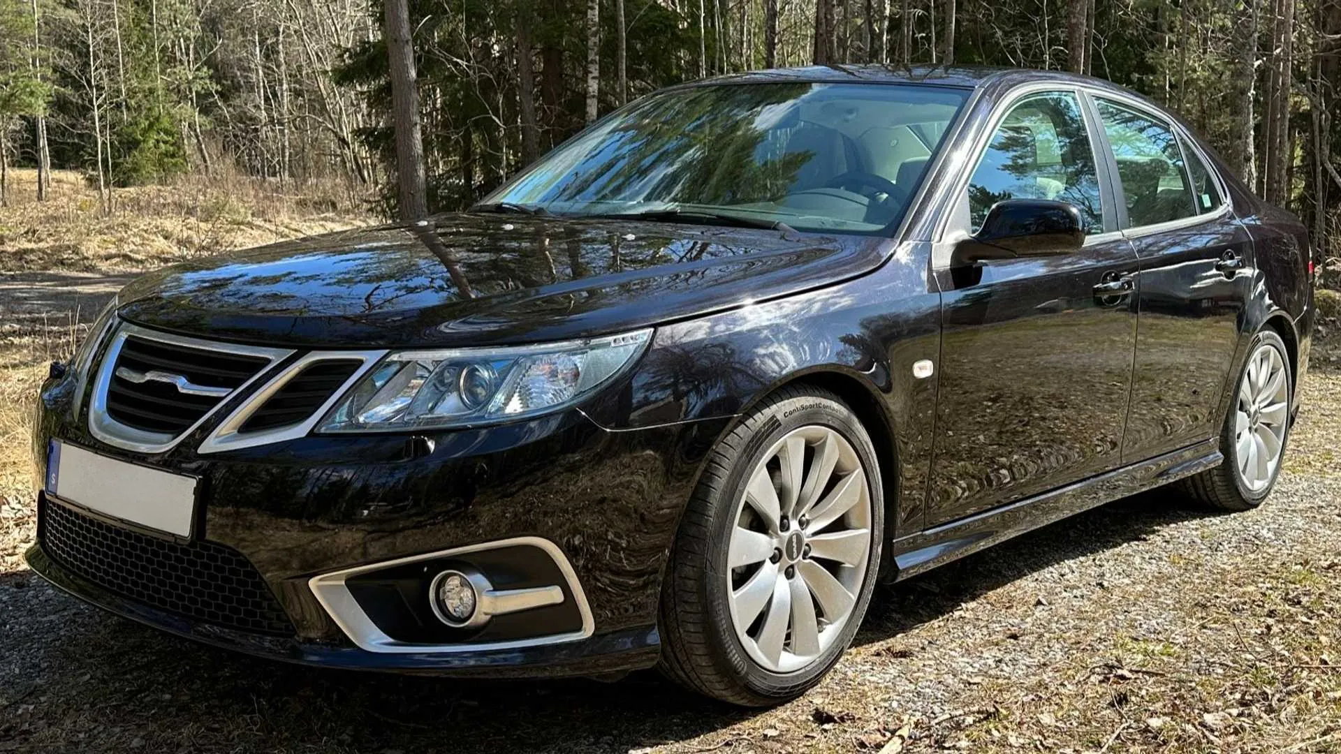 A glossy black Saab sedan is parked on a gravel road in a forest. The car is angled towards the viewer, showcasing its front grille, headlights, and side profile. The surrounding trees are bare, suggesting a cool season, and sunlight filters through the branches, casting reflections on the car's polished surface.