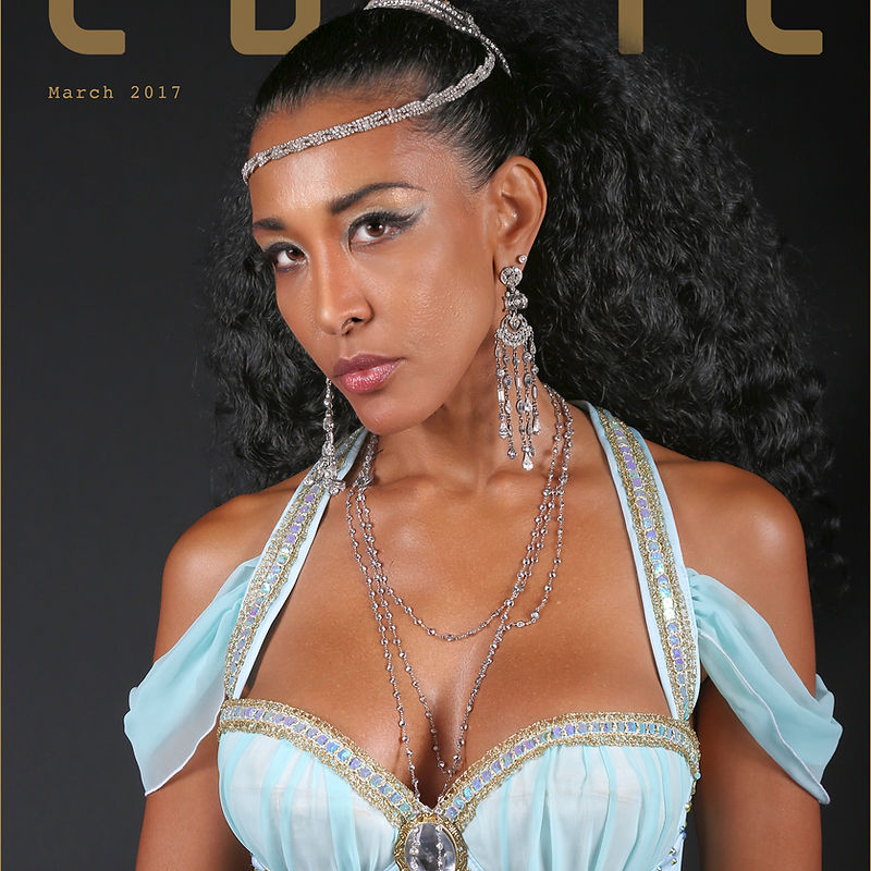 A striking portrait of a woman with dark curly hair and theatrical makeup, wearing a light blue, off-the-shoulder dress adorned with gold trim and intricate jewelry. She is wearing a jeweled headband and long, dangly earrings. The background is a dark, solid color, likely for a magazine cover or editorial shoot. The text 'March 2017' is visible in the upper left corner, and part of a large gold-lettered title is present at the top.