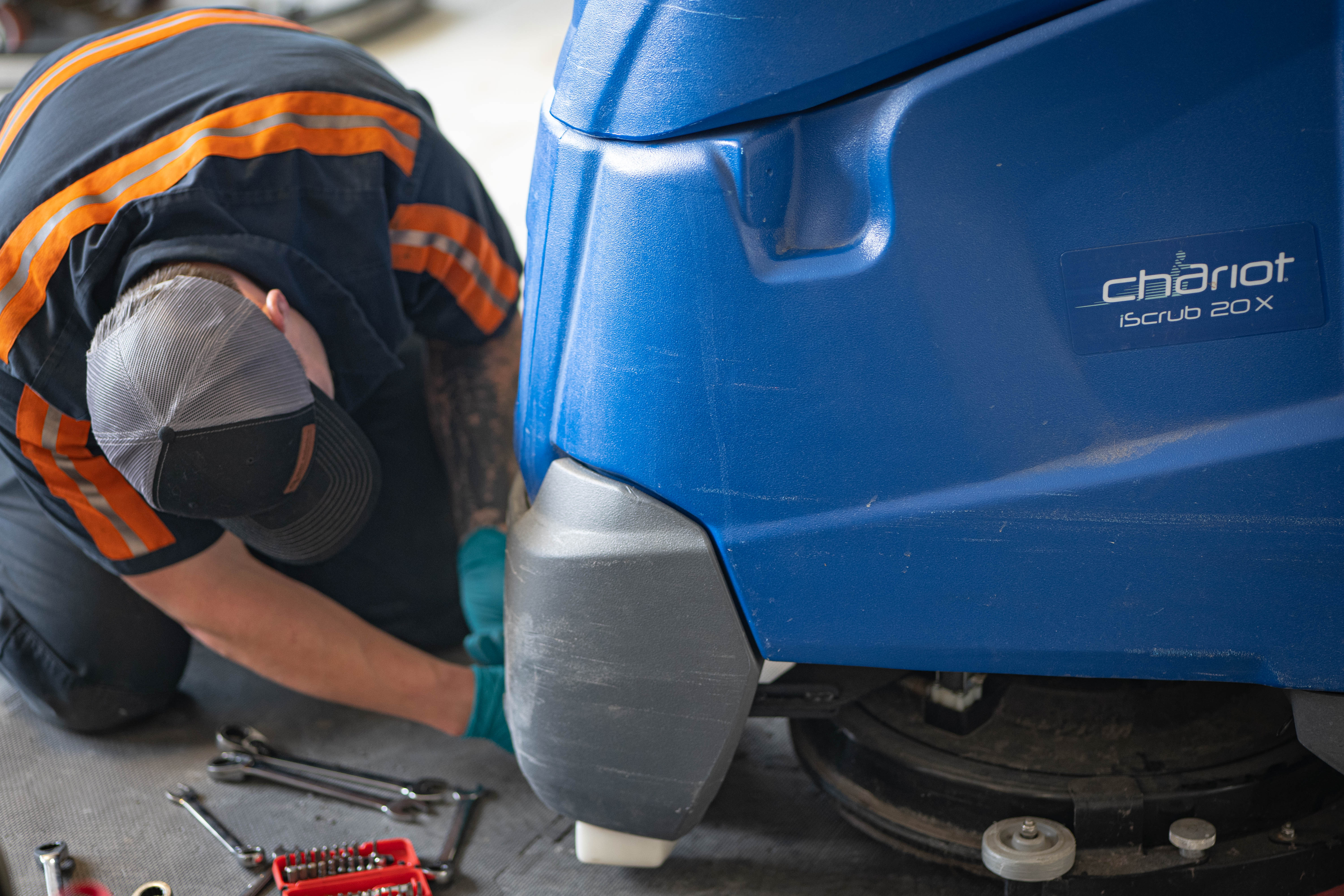 A close-up shot of a WaySolutions service specialist reaching underneath a blue Kärcher Windsor Chariot iScrub 20X stand-on floor scrubber to adjust the scrub deck or drive motor assembly. Wrenches and a socket kit are placed ready on the floor mat, illustrating deep mechanical preventative care on major supermarket cleaning assets.