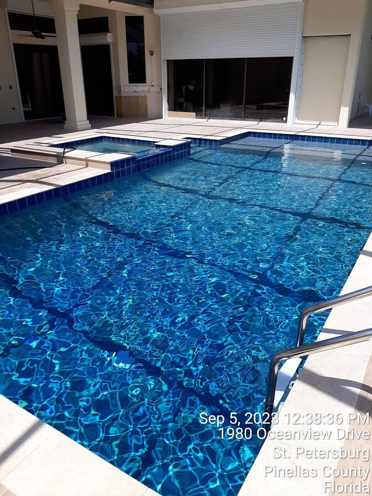 Rectangular residential swimming pool with deep blue water, decorative tile, and a covered patio area beside the pool.