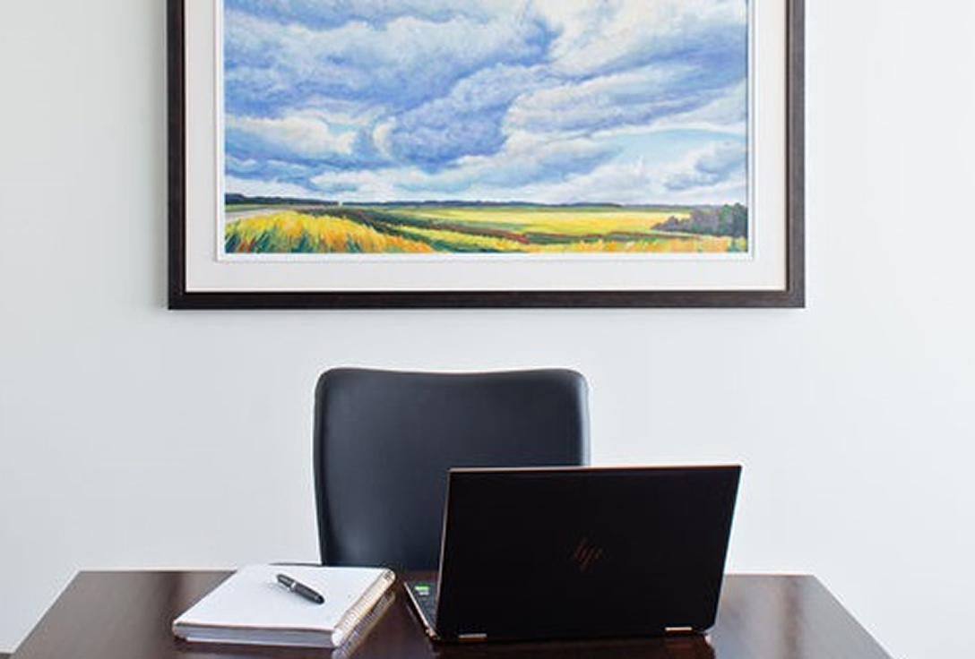 Office desk with a laptop and notebook. A large landscape painting of fields and sky hangs on a white wall, creating a calm and professional atmosphere.