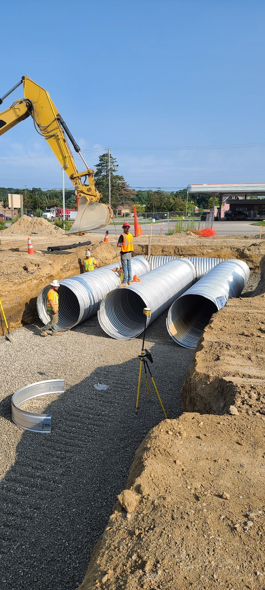 Construction workers install large corrugated metal pipes in a trench for stormwater drainage. An excavator assists placement while crews prepare aggregate bedding and verify alignment requirements.