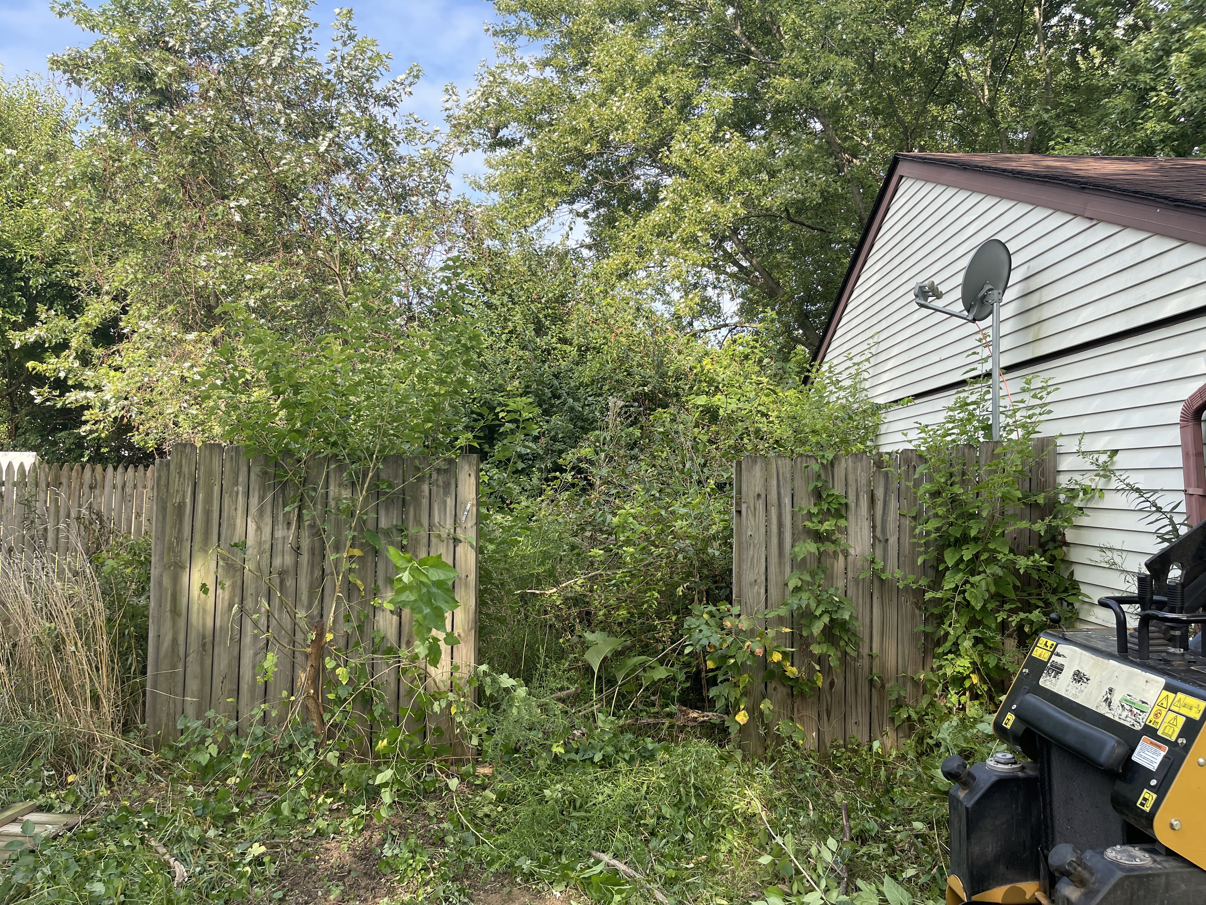 A backyard scene with an overgrown wooden fence, lush green trees, and the side of a house with a satellite dish. A piece of heavy machinery, possibly a stump grinder, is visible on the right side of the frame, with caution labels and fuel caps.