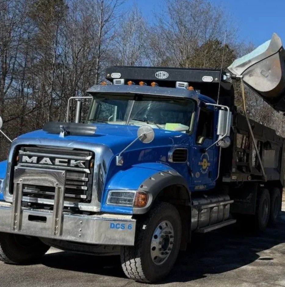 A blue Mack dump truck from Dinsmore Construction Services is parked outdoors with its dump bed raised. The heavy duty vehicle is positioned near trees at a worksite.