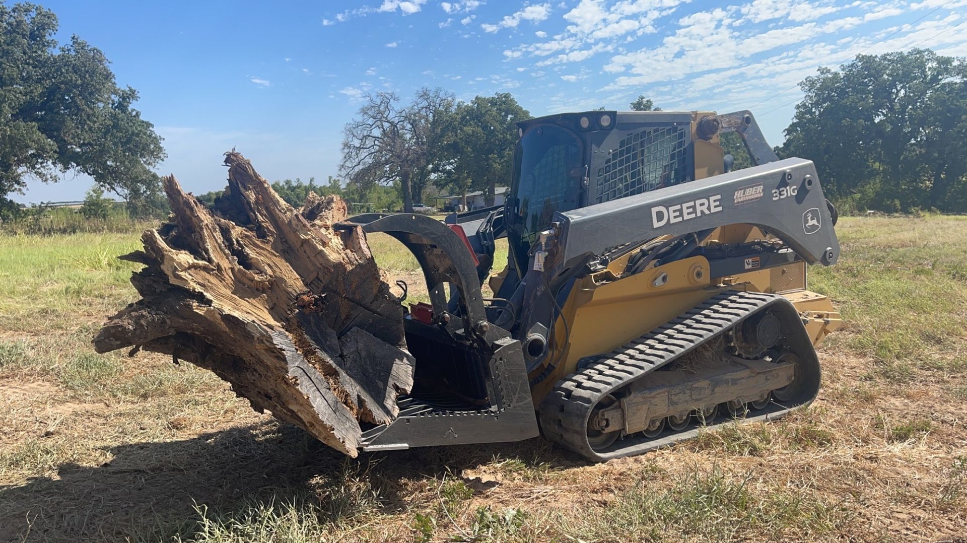 Skid steer lifting and transporting large tree stump using grapple attachment, demonstrating debris removal, land clearing, and heavy material handling services for site preparation and property development projects outdoors.