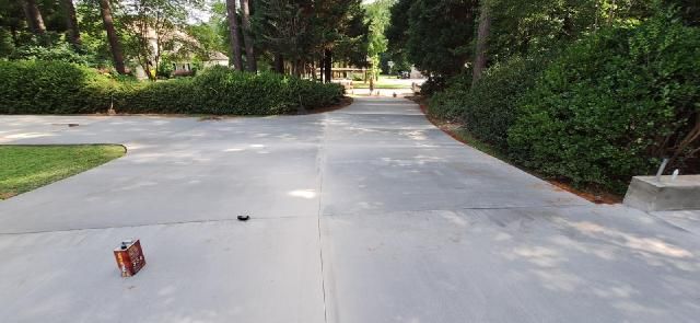 Long residential concrete driveway bordered by trimmed hedges and tall trees, smooth finished surface with expansion joints, leading toward street in quiet suburban neighborhood setting.