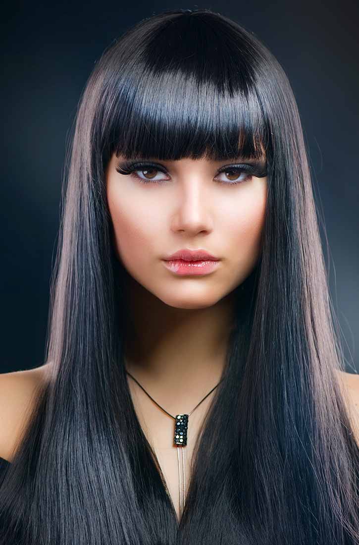 A close-up portrait of a woman with long, straight black hair and bangs. She is wearing dramatic false eyelashes, and her lips are parted slightly. A dark pendant with sparkling accents hangs from a thin necklace around her neck. The background is a dark, gradient shade.
