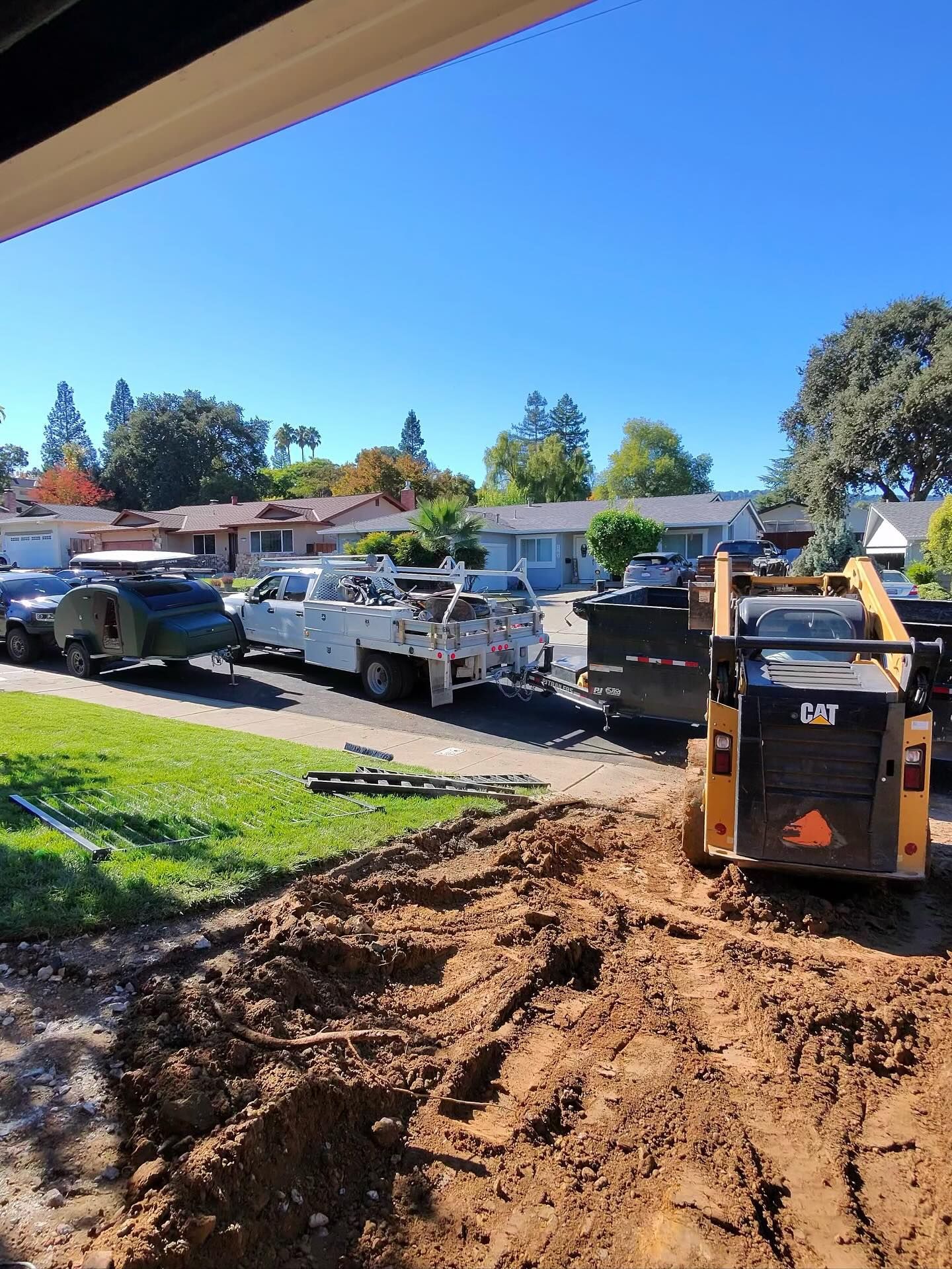 Residential construction site with skid steer loader grading soil, alongside parked work trucks and trailer, showcasing excavation, site preparation, and equipment staging in a suburban neighborhood under clear blue sky.