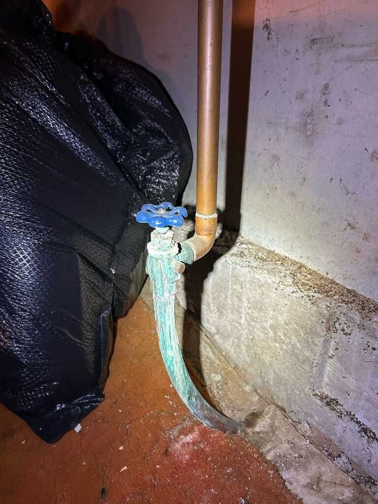 Aged copper plumbing pipe and blue handled shutoff valve show heavy green corrosion near a concrete  