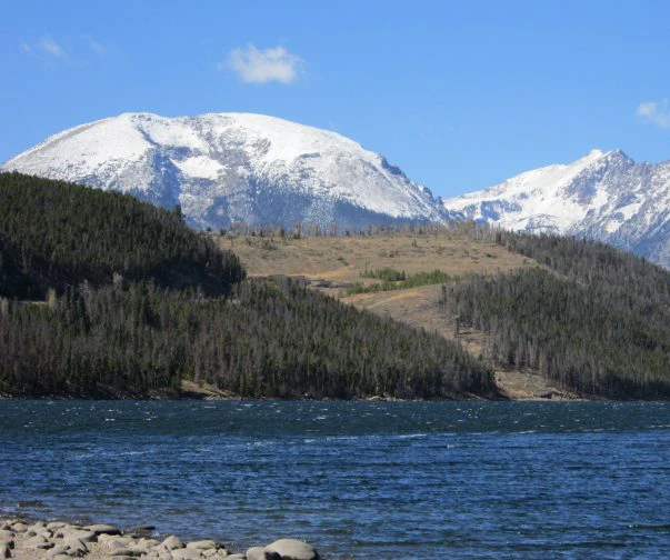 A serene view of a Colorado mountain range with a crystal-clear lake in the foreground. Snow-capped peaks rise in the distance, framed by dense forests and rocky terrain.