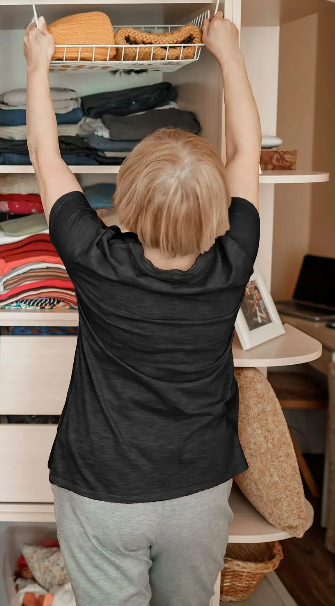 Rear view of a woman organizing a wooden closet, reaching up to hang a wire storage basket filled with folded yellow knits while neatly arranged folded clothing sits on the shelves below.