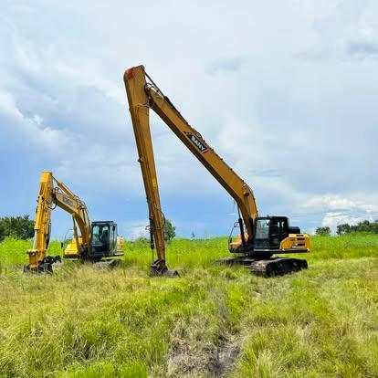 A Sany long-reach excavator and a smaller excavator parked in an overgrown grassy field under a cloudy sky.