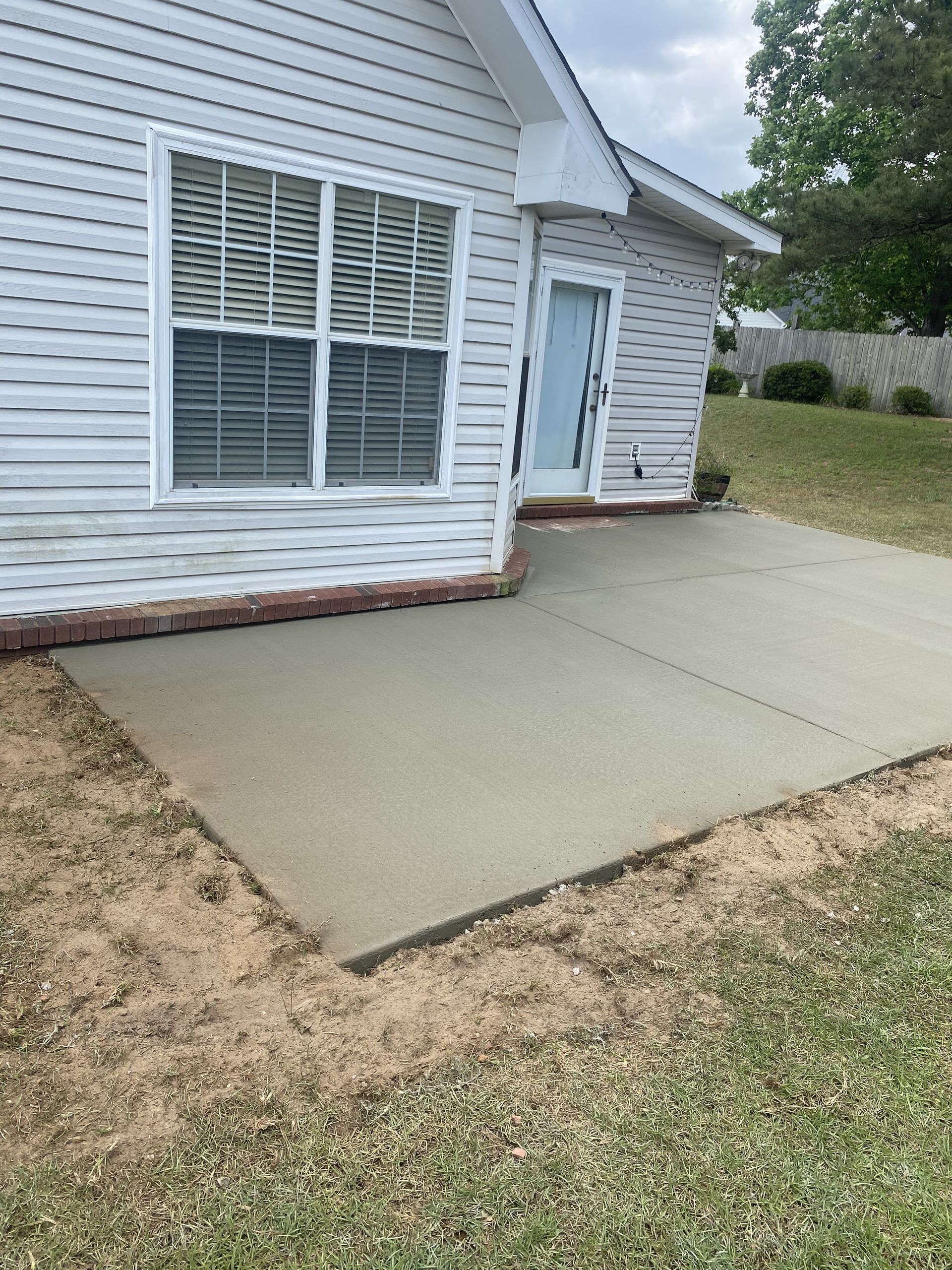 Newly poured concrete patio slab adjacent to residential home with vinyl siding, brick trim, large window, and back door, surrounded by grass and freshly graded soil edges.