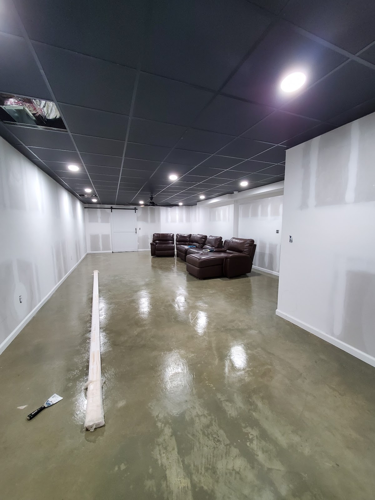 A room in what appears to be a basement or unfinished space, featuring a polished concrete floor, white walls with exposed drywall seams, and a dark grey drop ceiling with recessed lighting. A large sectional sofa made of dark brown leather is positioned in the corner of the room, and a sliding barn door is visible on the far wall. A long, wrapped roll of material and a putty knife are on the floor in the foreground.