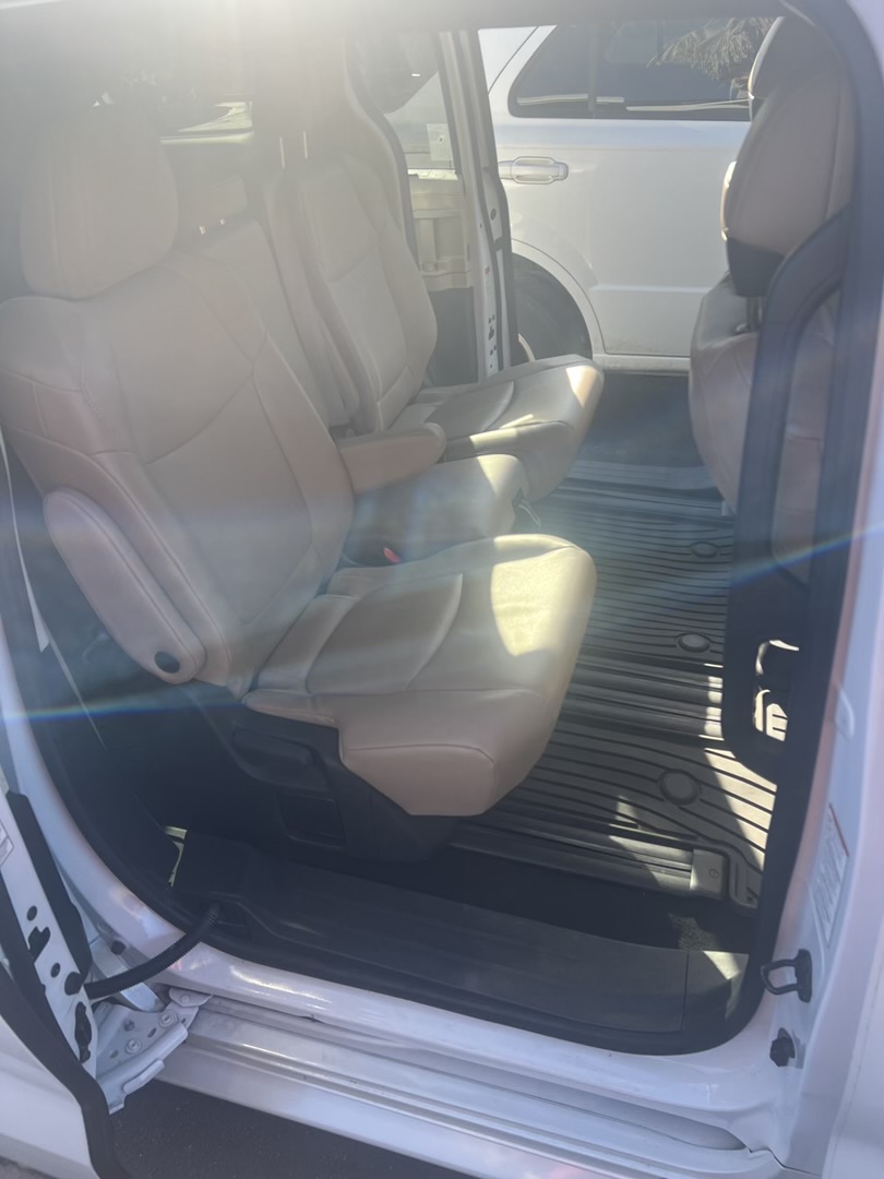 Interior of a white minivan with tan leather seats, illuminated by bright sunlight streaming through the windows. The front two rows of captain's chairs are visible, along with rubber floor mats. Another white vehicle is partially visible through the open side door.