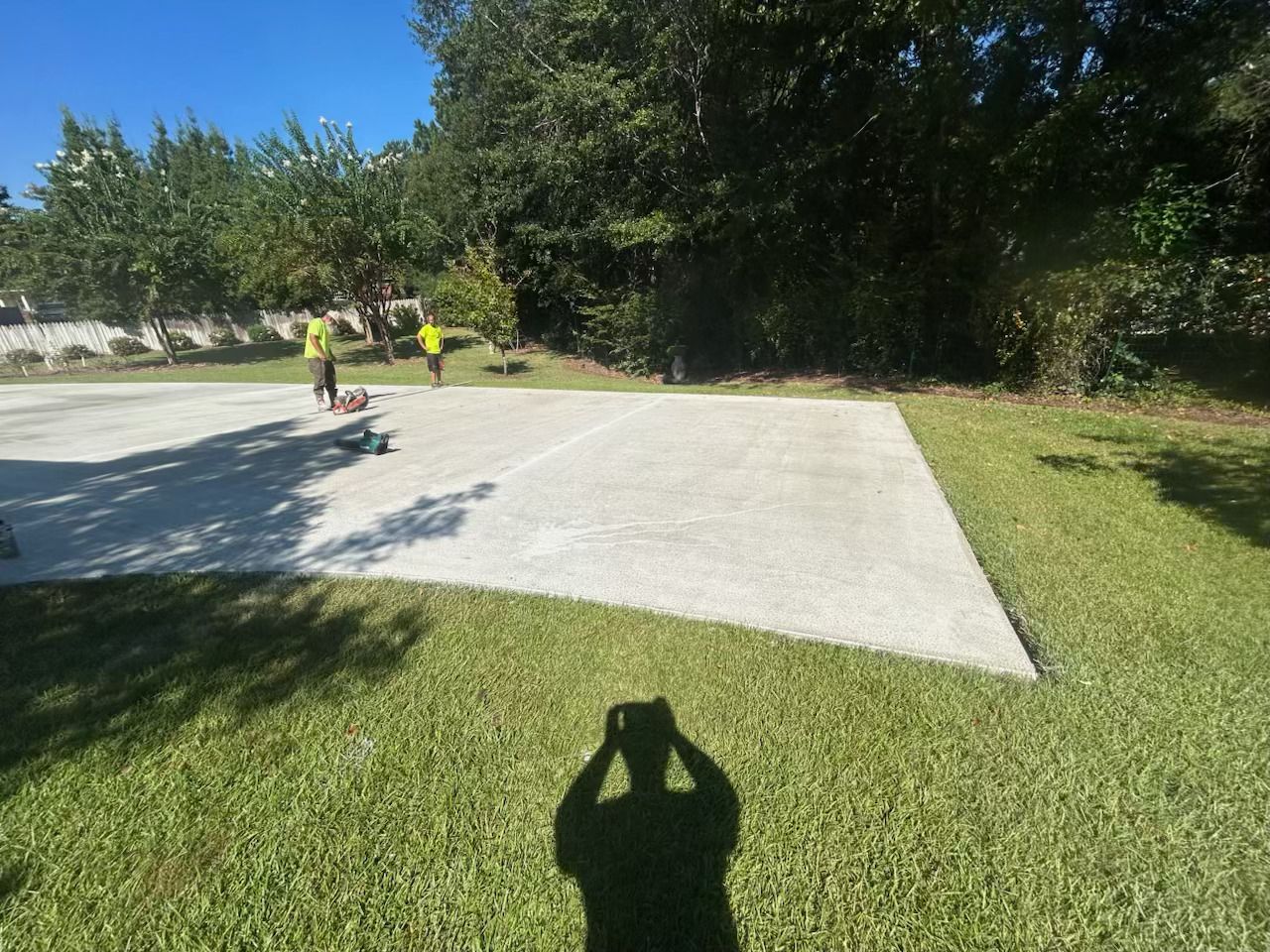 Two workers finishing large outdoor concrete slab in grassy backyard, using power tools on smooth surface, surrounded by trees and fence, under clear blue sky during daylight.