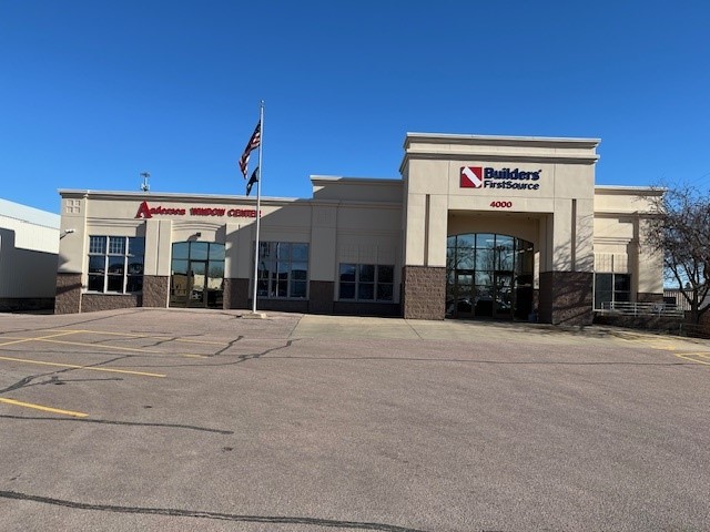A large beige building with a sign reading "Builders FirstSource" and the address "4000" is shown. The building also features a secondary sign for "Andersen Window Center." The structure has a grand entrance with tall windows and a mix of beige and brown tones on the facade. A flagpole with the U.S. and state flags stands prominently in the parking lot, which is mostly empty. The sky is clear and blue.